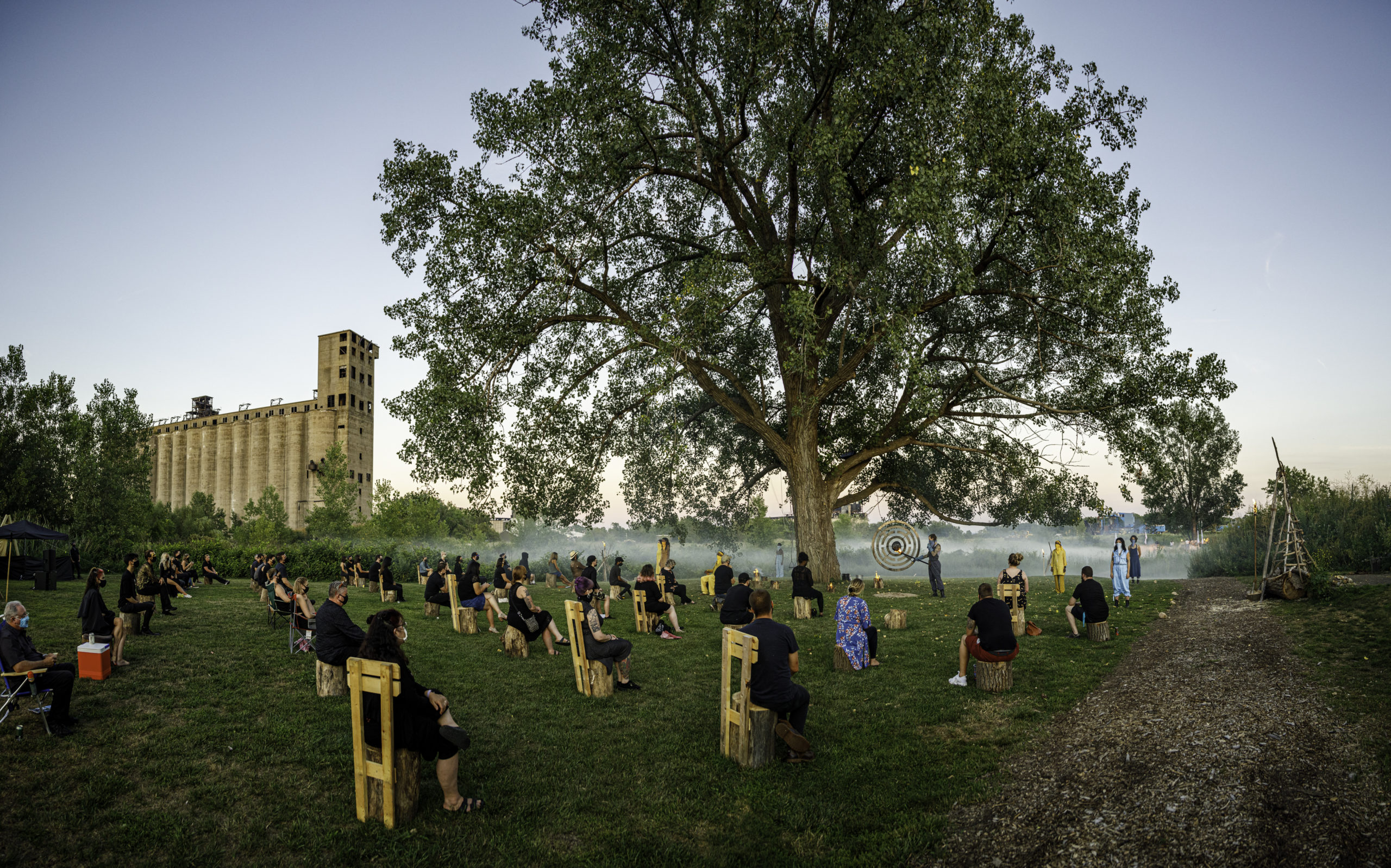 Photograph of a large tree in a grassy field with smoke blanketing the ground like mist, in the background the large imposing concrete grain elevator, with individual audience members seated in wooden chairs, each distanced from one another, around the tree.